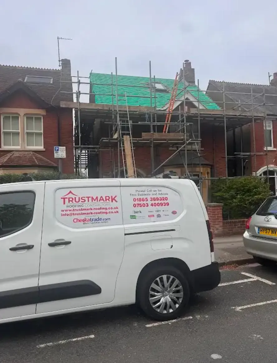 Trustmark Roofing company van with branding, parked on a street in front of a house with scaffolding and a new roof being installed, showing green underlayment.