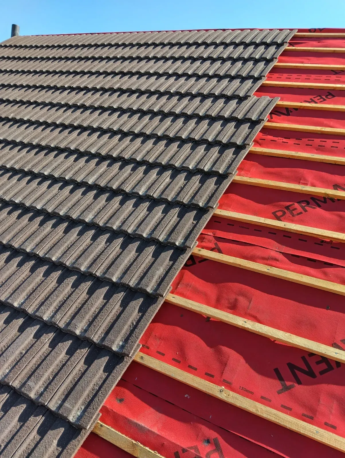 New concrete roof tiles being installed on a pitched roof over red breathable membrane and wooden battens.
