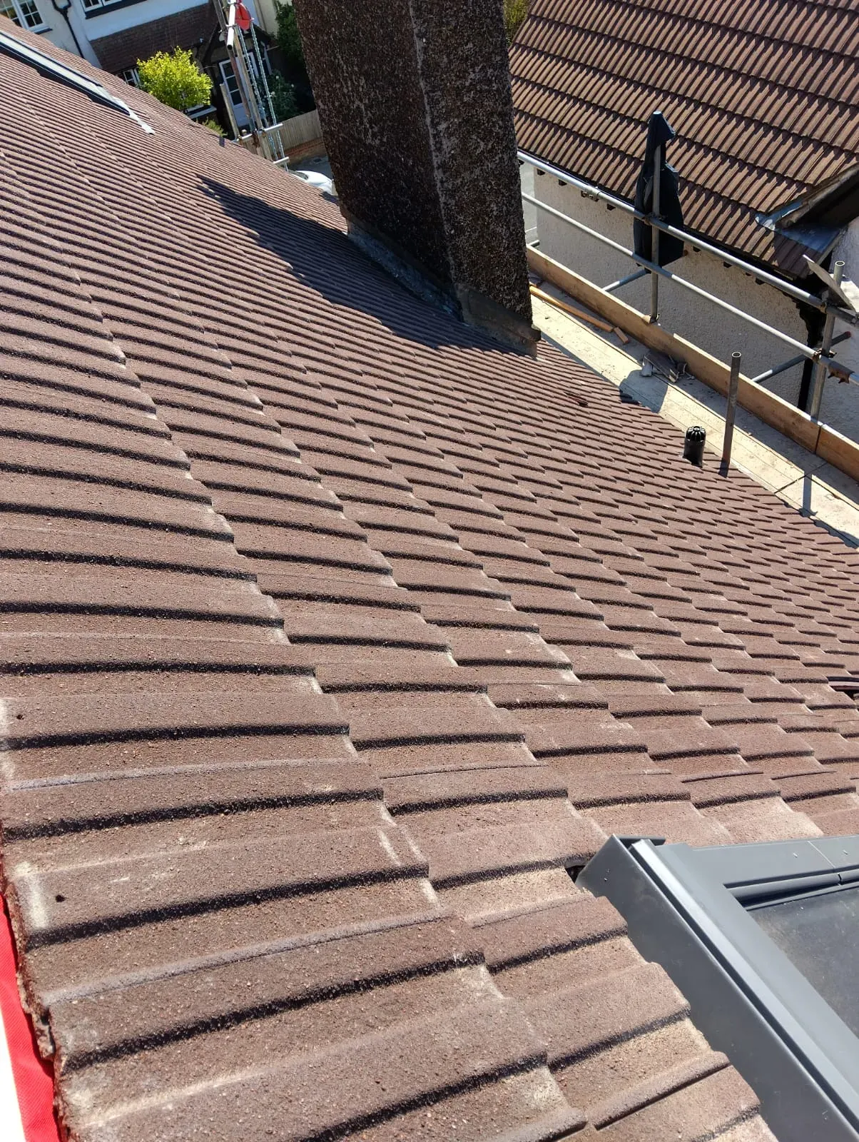 Close-up of a pitched roof with new brown interlocking concrete tiles, a chimney, and a modern roof window installation, with scaffolding nearby.