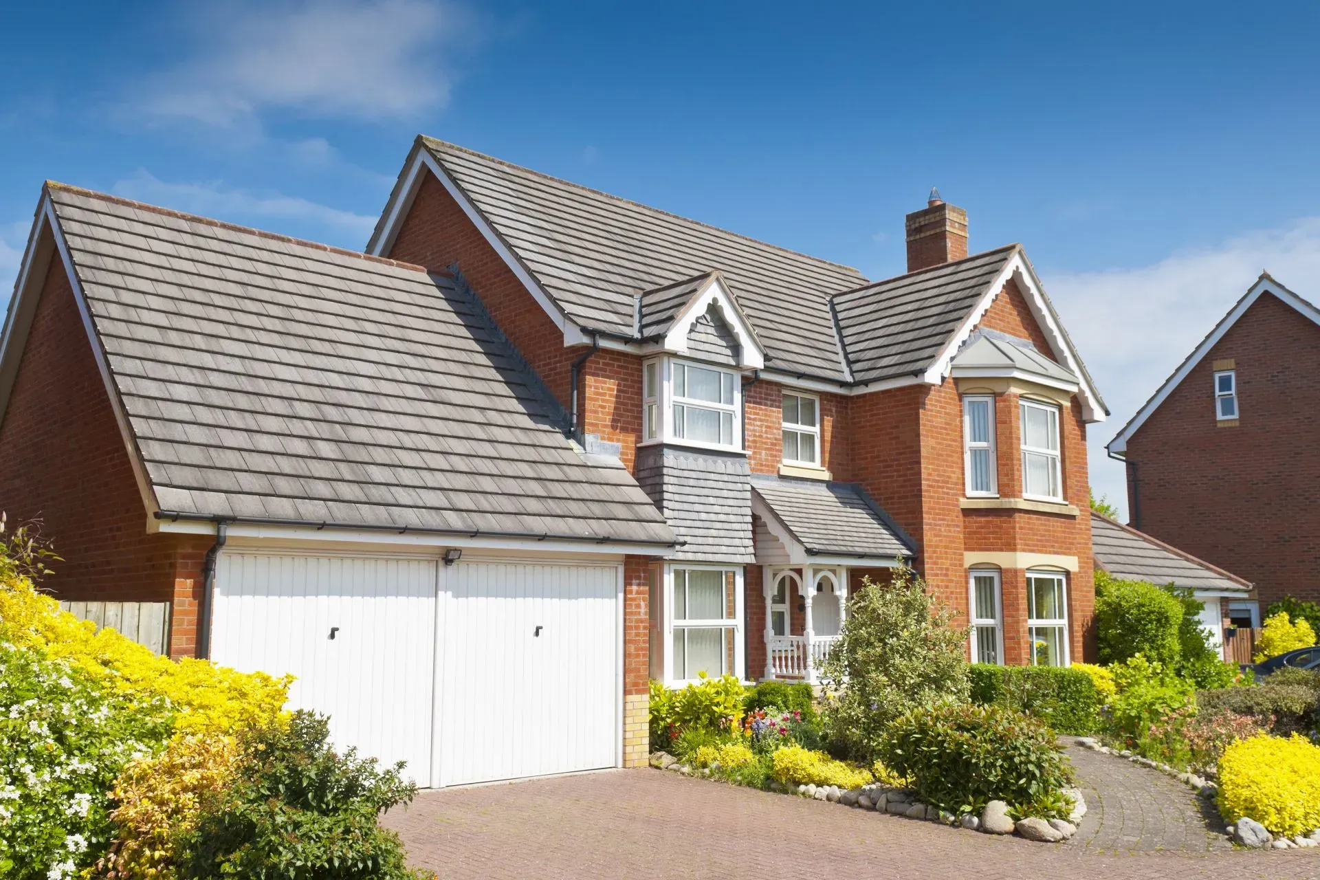 A modern red brick house with a grey tiled roof and white garage doors, surrounded by a landscaped garden.