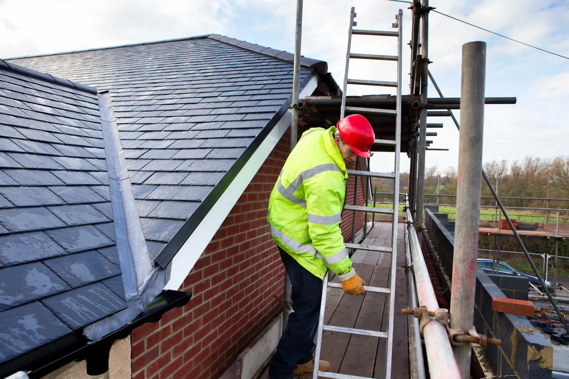 Professional roofer in a high-vis jacket and red hard hat on scaffolding, inspecting or working near a recently installed slate or tiled roof on a new build.